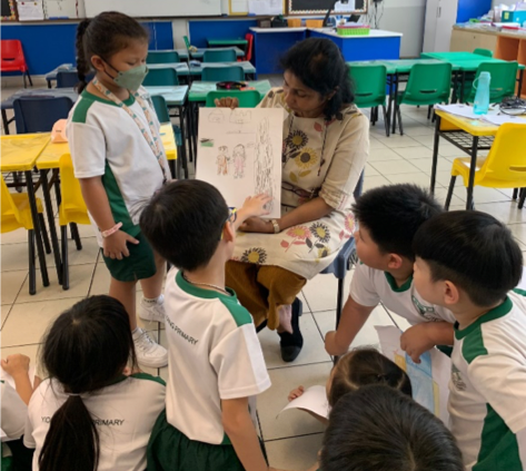 A teacher sits and shows a children's drawing to a group of young students in white and green uniforms in a classroom.